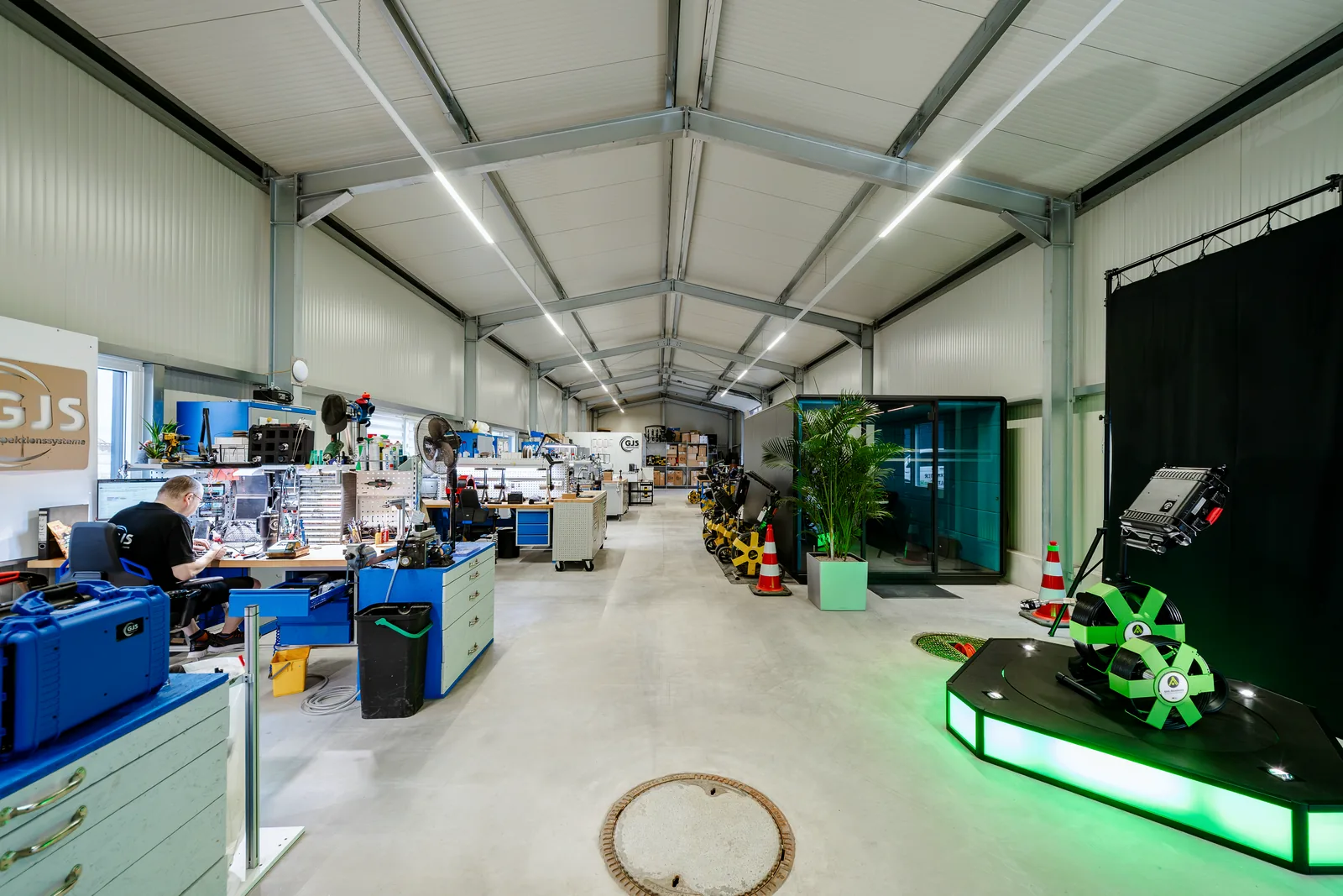Firmenhalle mit Mitarbeiter an der Werkbank, Schachtdeckel im Boden und dem leuchtenden Podest mit dem SAG-Kamerasystem